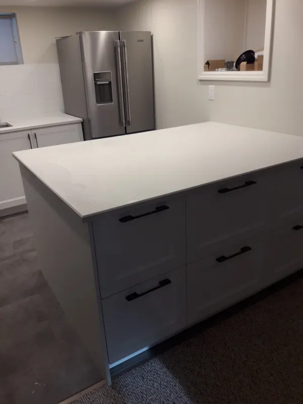 Kitchen island with a white quartz top and white shaker drawers with black pulls, beside a stainless French-door fridge